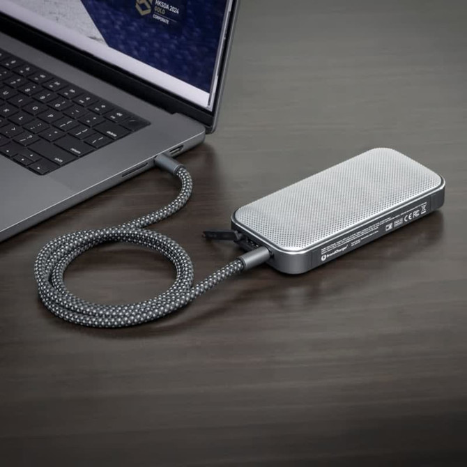 A silver Bluetooth speaker power bank with a textured surface, connected to a laptop with a braided cable.