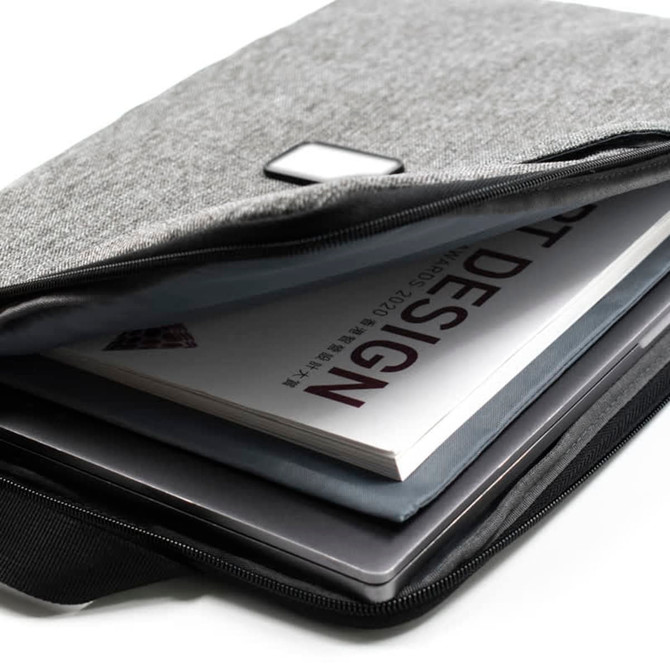 A grey laptop sling bag with a zip, containing a laptop and a design book. It features a small logo.