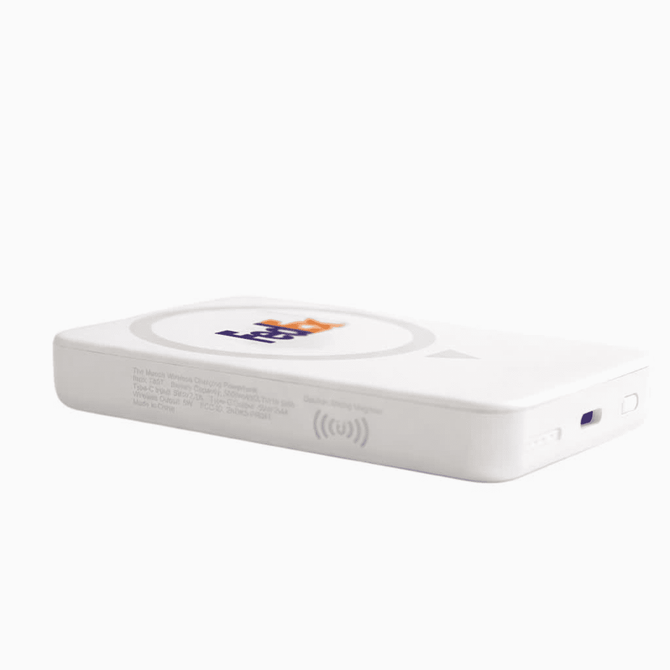 A white wireless charging power bank with a logo, resting on a desk near plants and a laptop.