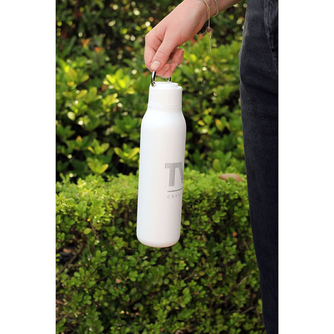 A drink bottle with a metal loop, featuring a white finish and a logo, held in a person's hand against greenery.
