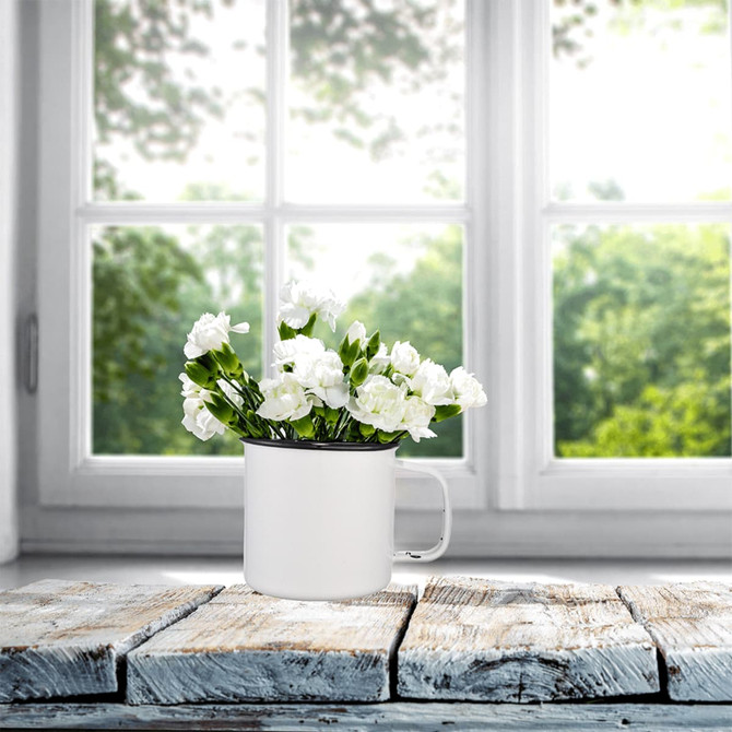 A white enamel metal cup filled with white flowers, placed on a rustic wooden surface near a sunny window.