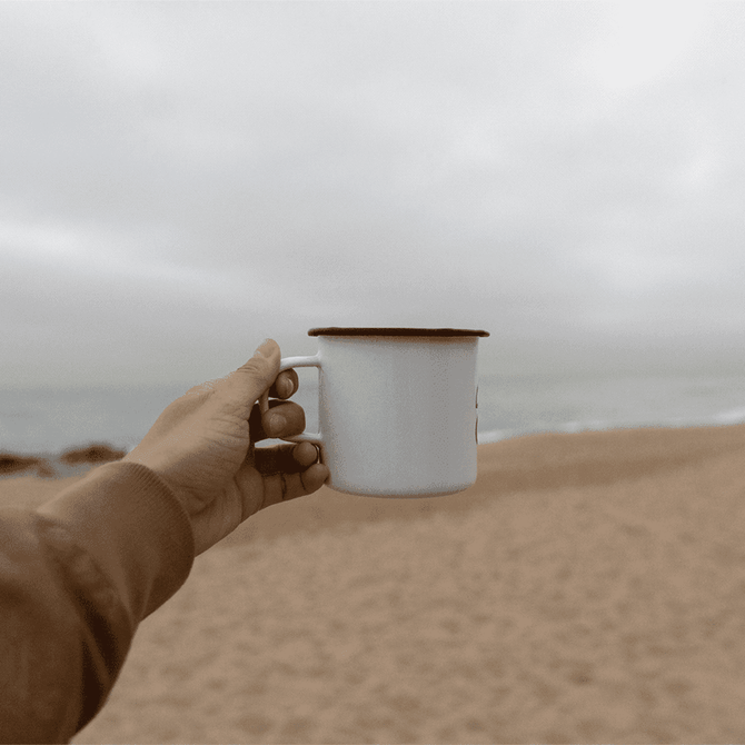A drink bottle featuring a white enamel metal cup with a black rim, held against a beach backdrop.