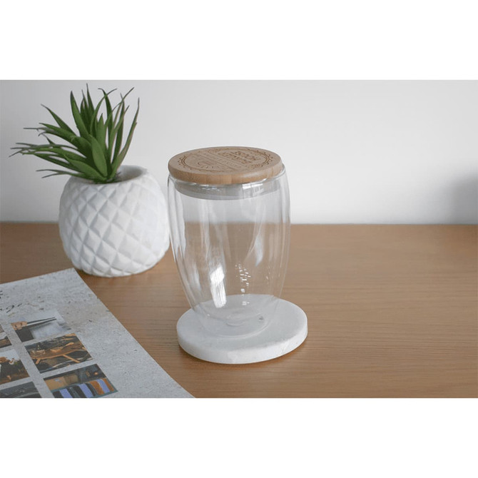 A drink bottle made of clear glass with a bamboo lid, set on a white marble base, alongside a decorative plant.