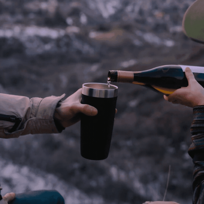 A drink bottle with a stainless steel interior, featuring a black exterior, being filled from a wine bottle outdoors.