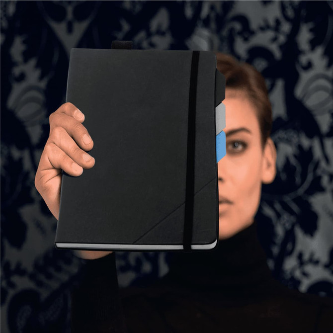 A black notebook held by a person, featuring a coloured tab and elastic strap, against a patterned background.