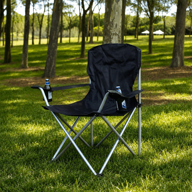 An oversized folding chair in black with silver accents, set on green grass in a wooded area.