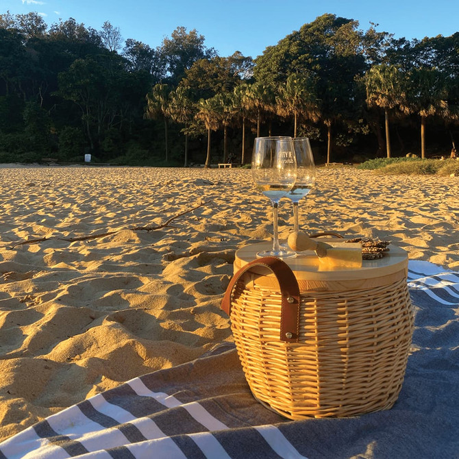A wicker cooler basket in natural colour with a leather strap, resting on a beach blanket, with a glass of wine beside it.