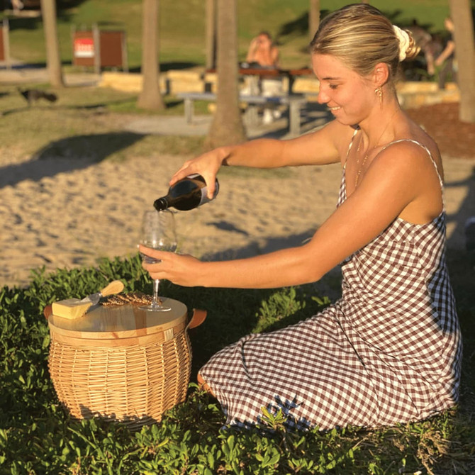 A wicker cooler basket in a natural finish, featuring a woman pouring a drink into a glass beside it.