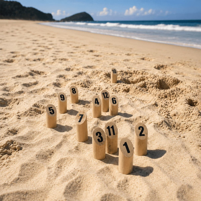 A set of wooden skittle pins numbered 1 to 12 arranged in sand on a beach, with a scenic ocean backdrop.
