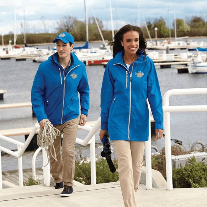 A blue rain jacket with a hood, featuring a logo, worn by a man and a woman by a marina.