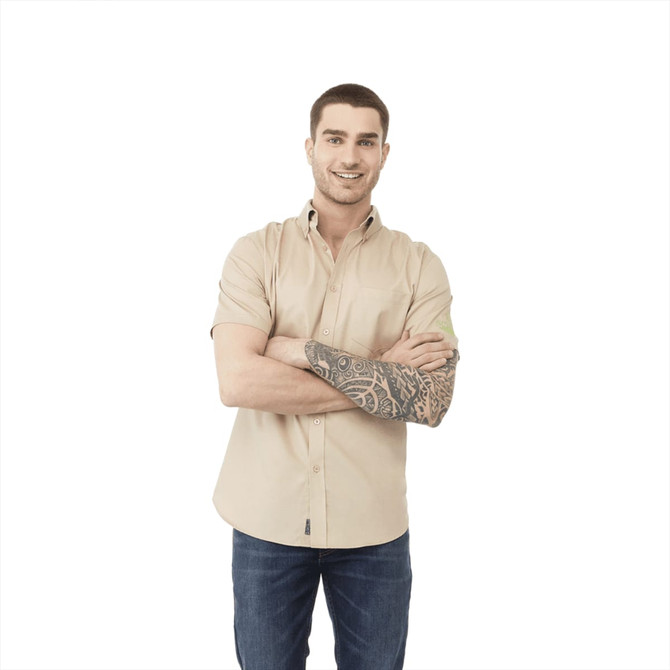 A man wearing a beige short sleeve button-up shirt with a tattoo, standing with arms crossed. The shirt has a logo.