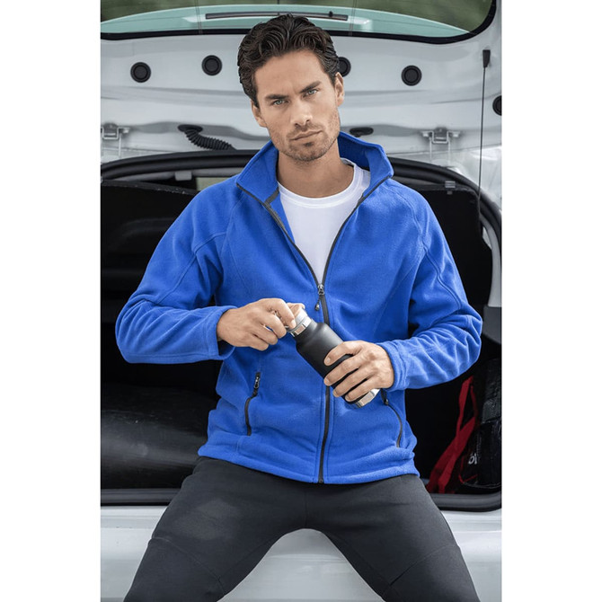 A man in a blue polar fleece jacket holds a drink bottle, seated in the back of a vehicle. The jacket has a logo.