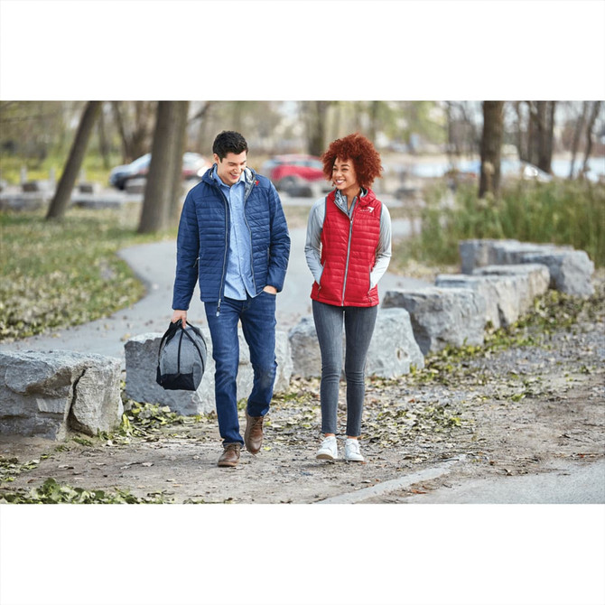 A man in a navy puffer jacket and a woman in a red puffer vest walk on a path, accompanied by a bag.