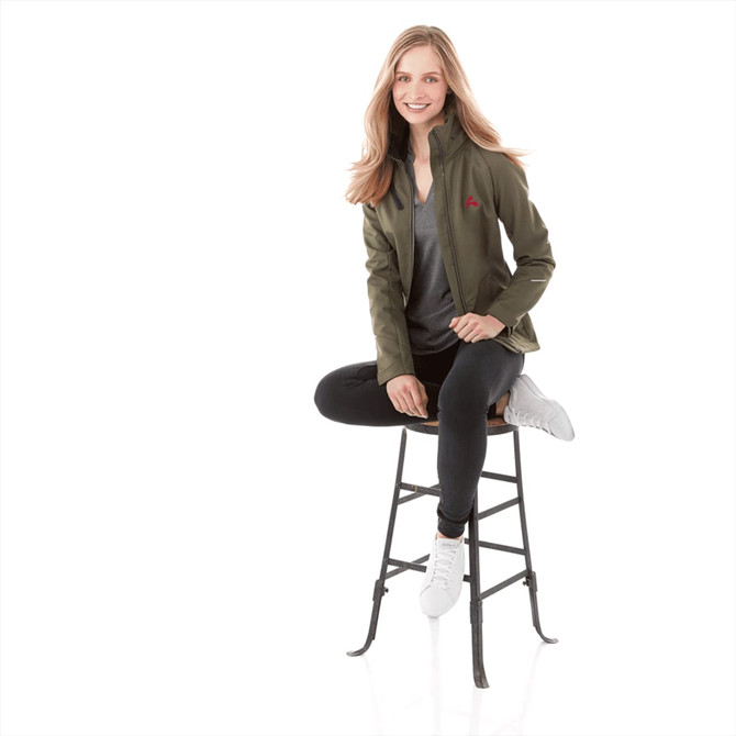A woman wearing a green Peyto softshell jacket, seated on a stool, styled with black pants and white shoes.