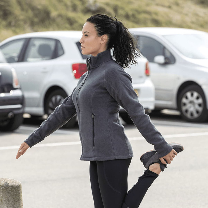 A woman stretches while wearing a grey polar fleece jacket, with parked cars in the background. The jacket features a logo.