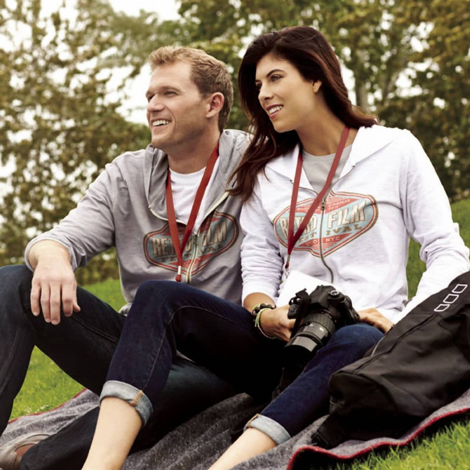 A grey and white zip-up hoodie worn by a woman and a man, both sitting outdoors with a camera. They have lanyards.
