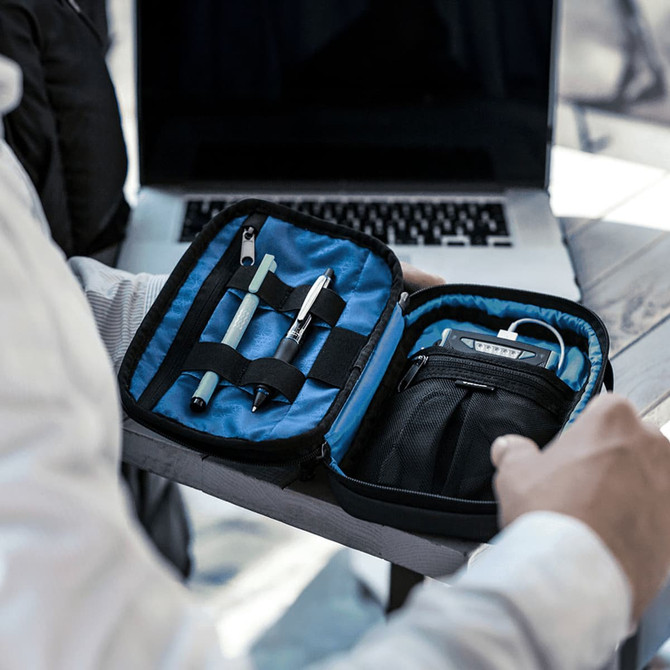 A black toiletry bag with a blue interior, containing pens and a small gadget, resting on a table beside a laptop.