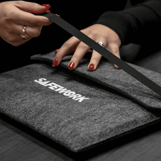 A grey felt laptop sleeve is being held, featuring a strap closure and a logo on the front.