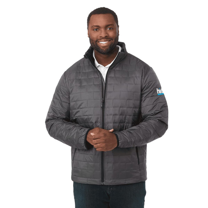 A man wearing a grey puffer jacket with a zip front and a logo on the sleeve.