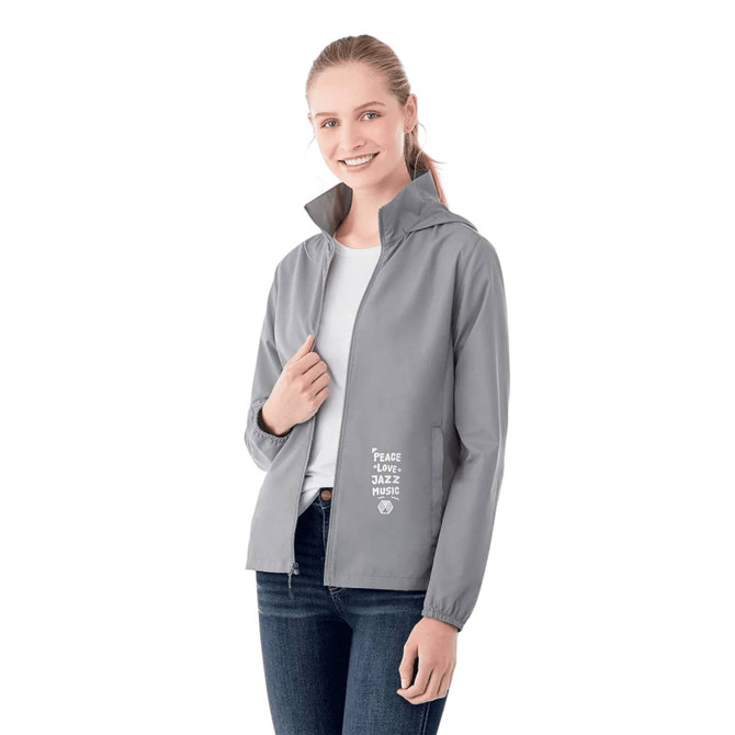 A woman wearing a grey packable rain jacket with a hood, featuring a logo and text on the front.