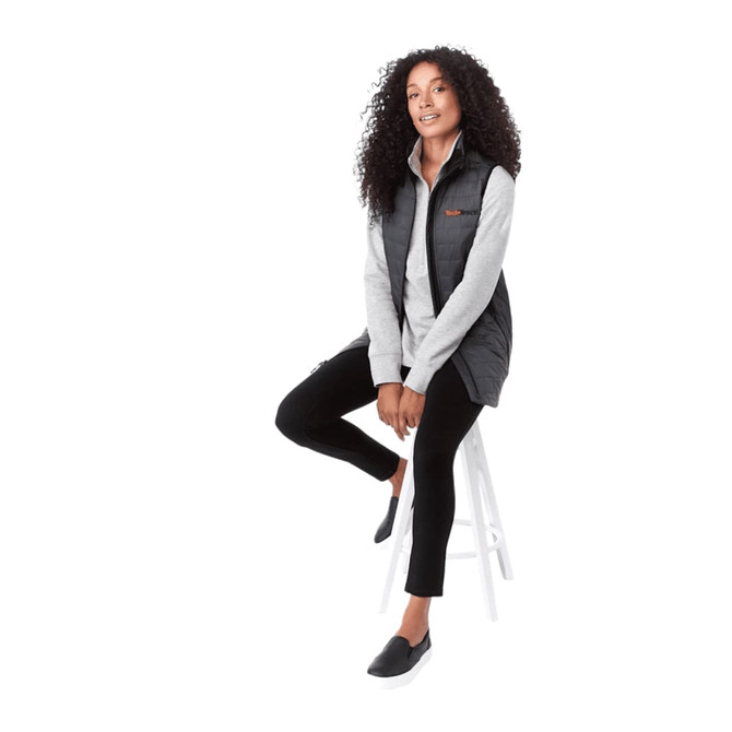 A woman sits on a stool wearing a black insulated puffer vest over a light grey top, paired with black trousers.