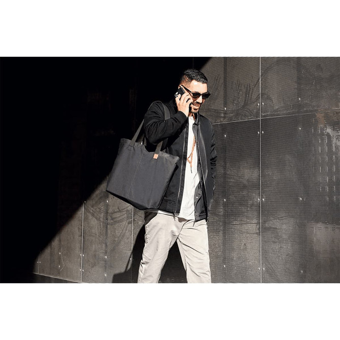 A person carrying a black tote bag, wearing sunglasses and a casual outfit, walking in a modern urban setting.