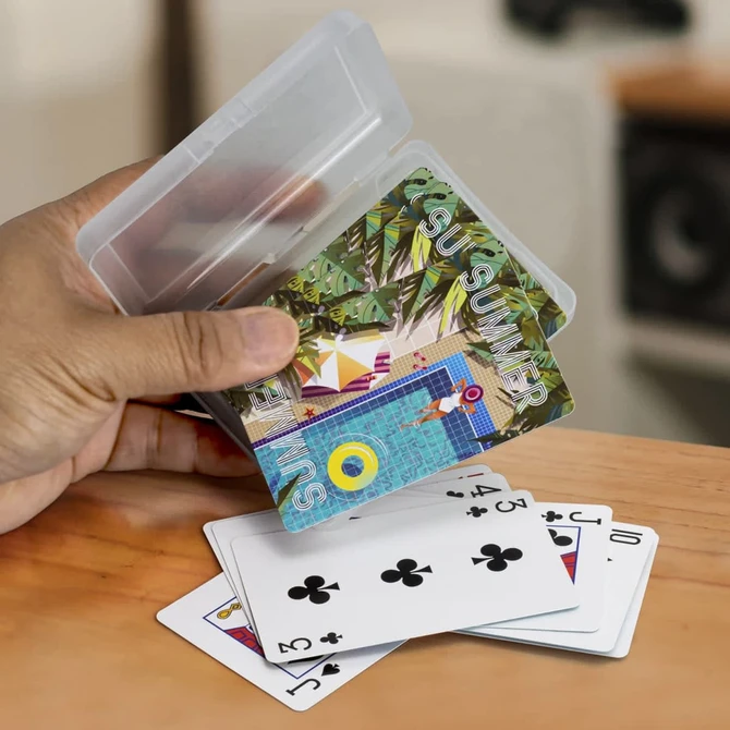 A hand holding a transparent case containing colourful playing cards, with some cards laid out on a table.