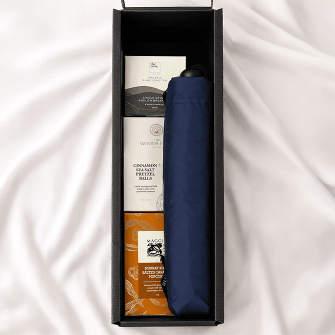 A navy umbrella is nestled in a box with treats like cinnamon balls and other snacks. The box has a sleek design and logo.