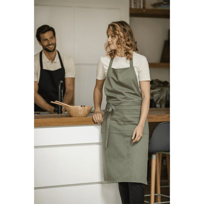 A green recycled apron is displayed on a woman standing by a kitchen counter, with a man in an apron in the background.