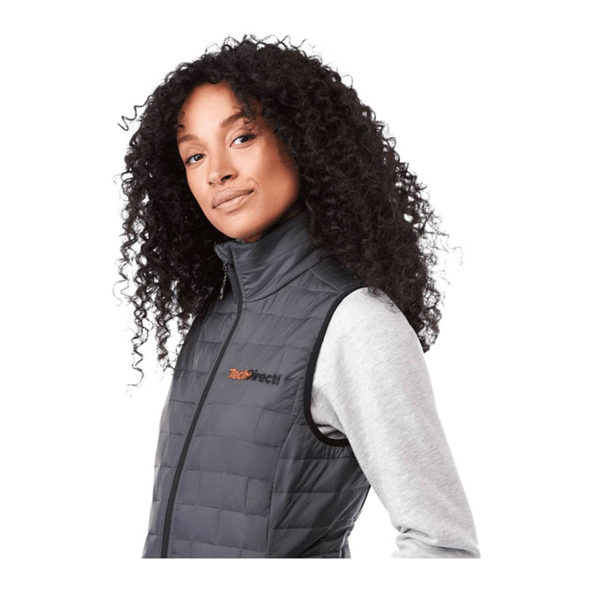 A woman with curly hair wears a black insulated puffer vest, featuring a logo on the chest.
