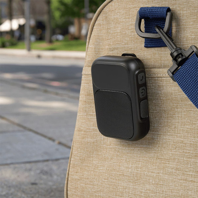 A black Bluetooth speaker attached to a bag, set against an outdoor urban background. It features controls and a logo.
