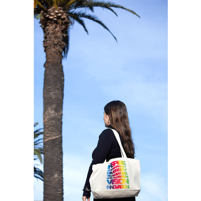 A person holding a multi-coloured zippered cotton canvas tote bag stands near a palm tree against a blue sky.