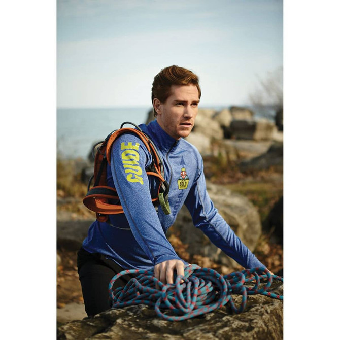 A man in a blue quarter zip jumper holds climbing ropes with a rocky landscape and lake in the background.