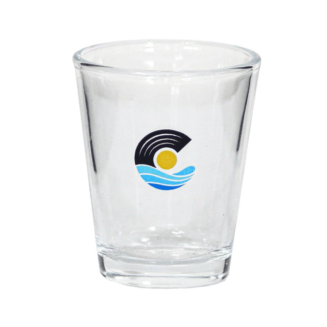A clear glass shot glass featuring a logo with blue waves and a sun design.