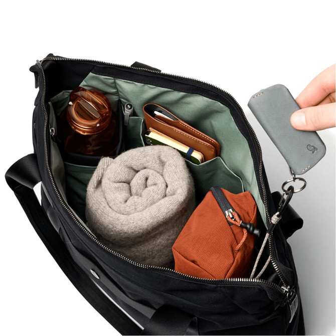 A black tote bag with a green interior, containing a drink bottle, a rolled blanket, a notebook, and a small brown pouch.