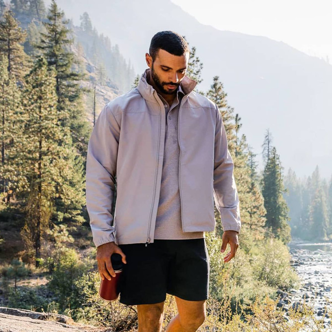 A full zip jacket in light grey, worn by a man outdoors, surrounded by trees and mountains. The jacket has a logo.