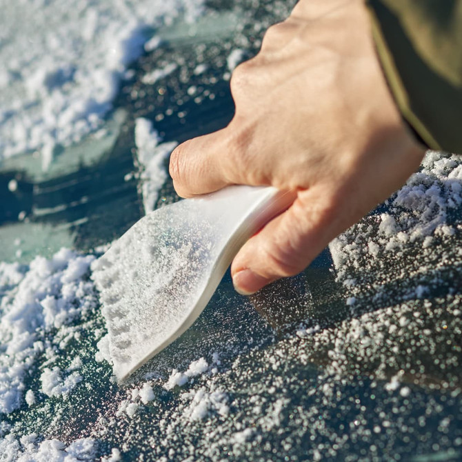 A white ice scraper being used to remove snow from a car windscreen, with a gloved hand gripping the scraper.