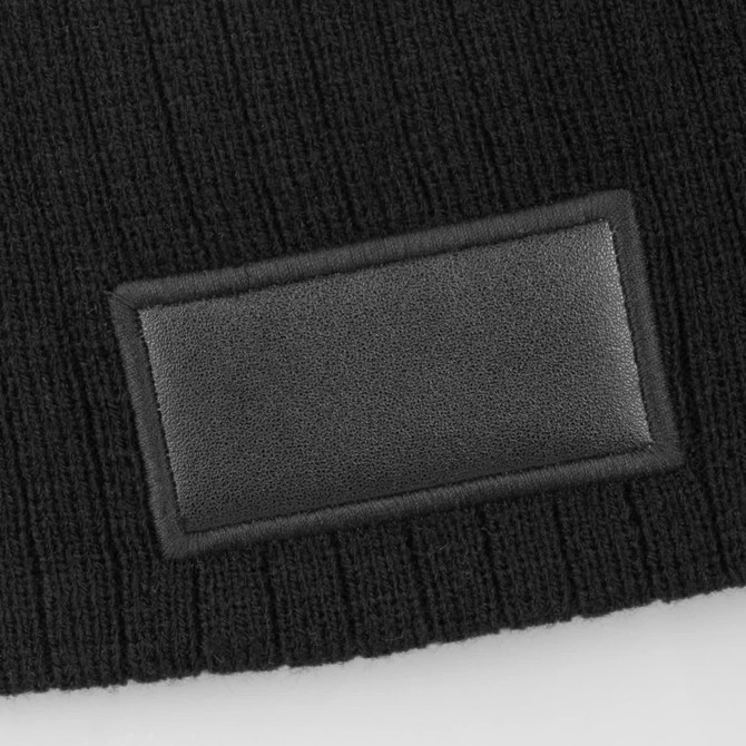 A black cable knit beanie featuring a textured patch. The material has a cosy, knitted appearance.