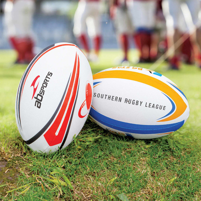 Two rugby league balls, one white with red accents and the other white with blue and yellow designs, on grass.