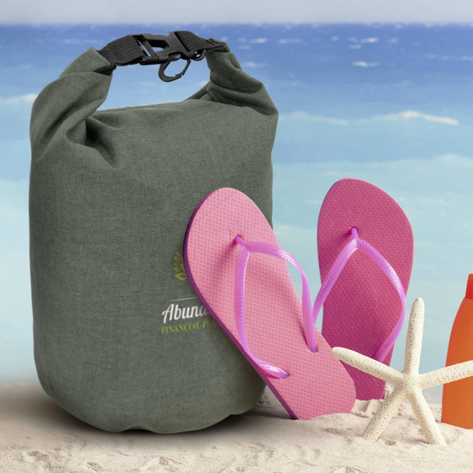 A green dry bag with a black clip sits next to pink flip-flops on a sandy beach background.