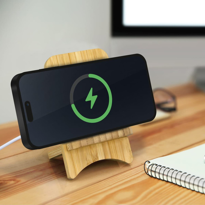 A bamboo wireless charging stand with a smartphone on it, displaying a charging icon. The stand is set on a wooden desk.