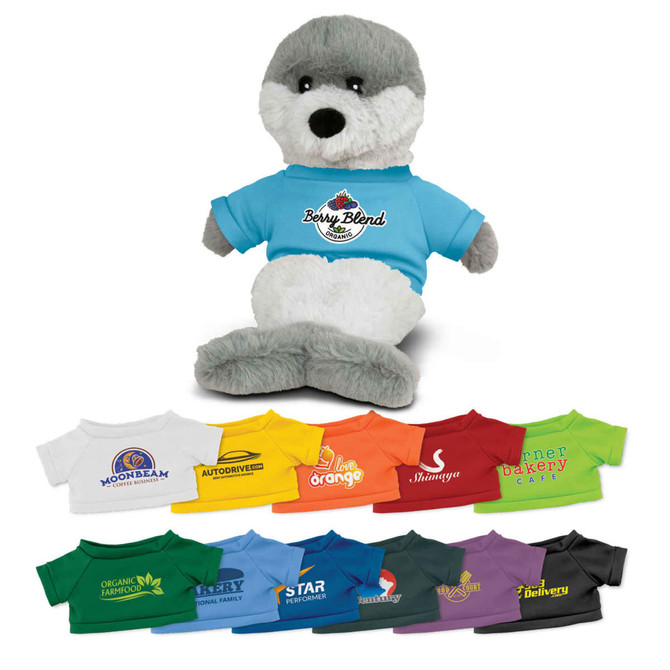 A grey and white seal plush toy wearing a blue t-shirt with a logo, alongside various t-shirts in different colours.