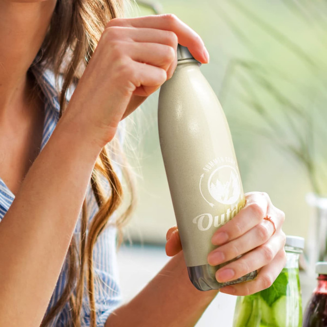 A drink bottle with a sleek, natural finish, featuring a logo and held by a person with wavy hair.