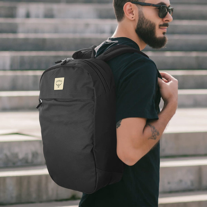 A black day backpack worn on the back of a person standing on steps, featuring a logo on the front.