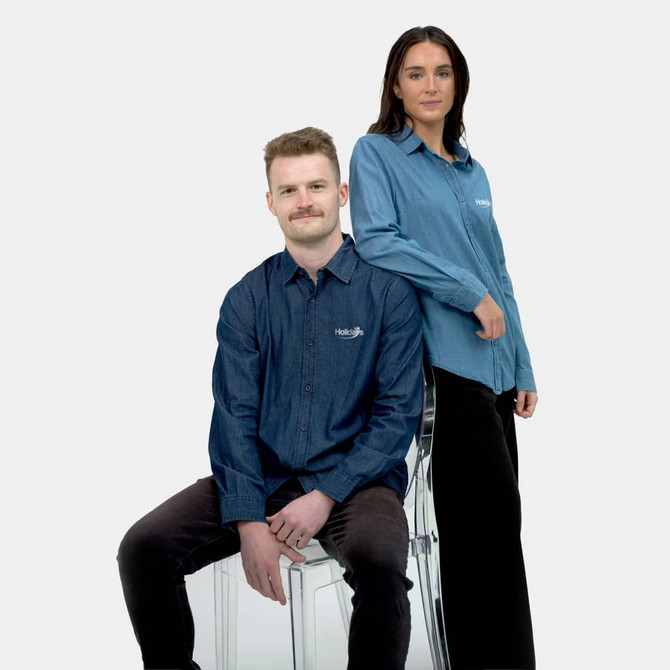 A denim shirt in two shades of blue, worn by a man sitting and a woman standing. The shirts feature a logo.