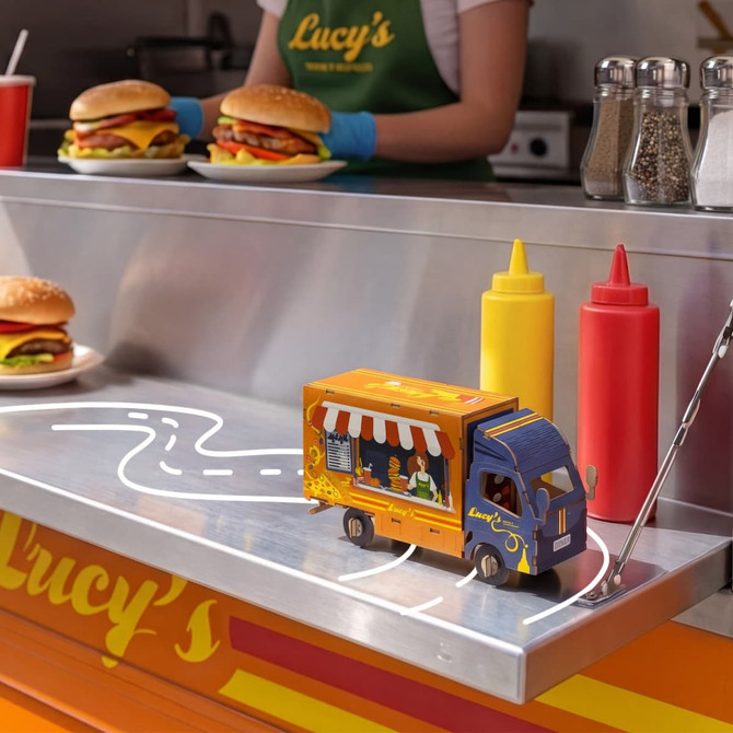 A wooden model of a small food truck in blue and orange, set beside condiments and burgers on a counter.