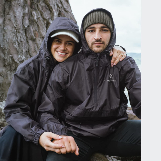 A couple wearing black windbreakers, seated outdoors near a tree, with one jacket featuring a logo.