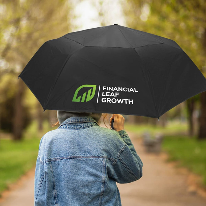 A black umbrella with a logo, held by a person wearing a denim jacket, on a tree-lined path.