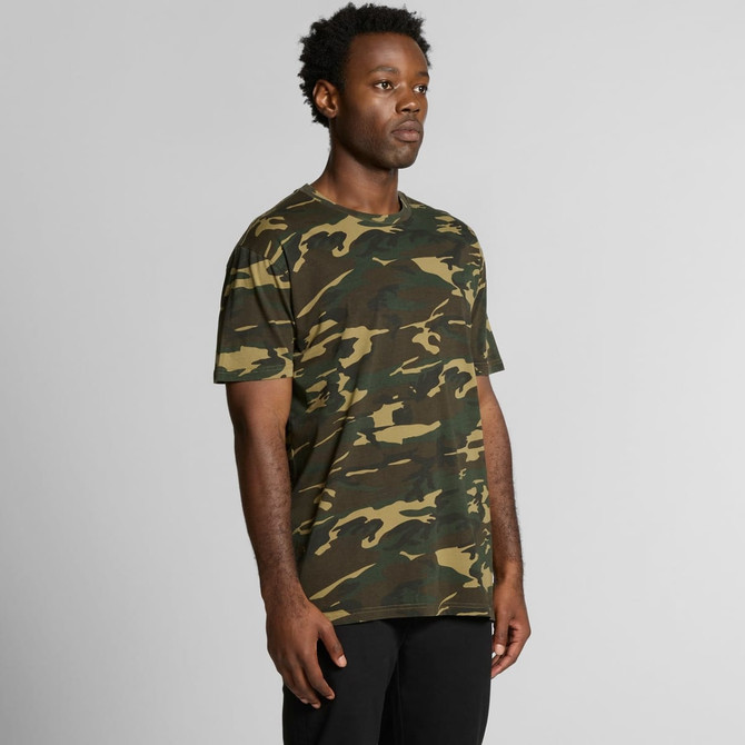 A man stands wearing a green and brown camo T-shirt, featuring a logo on the front. The shirt has a relaxed fit.