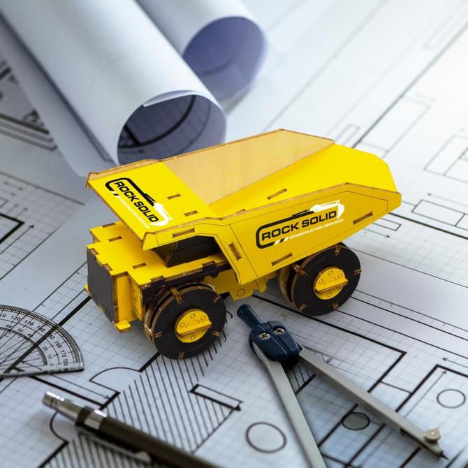 A wooden model of a yellow mining truck, placed on architectural blueprints with drafting tools beside it.